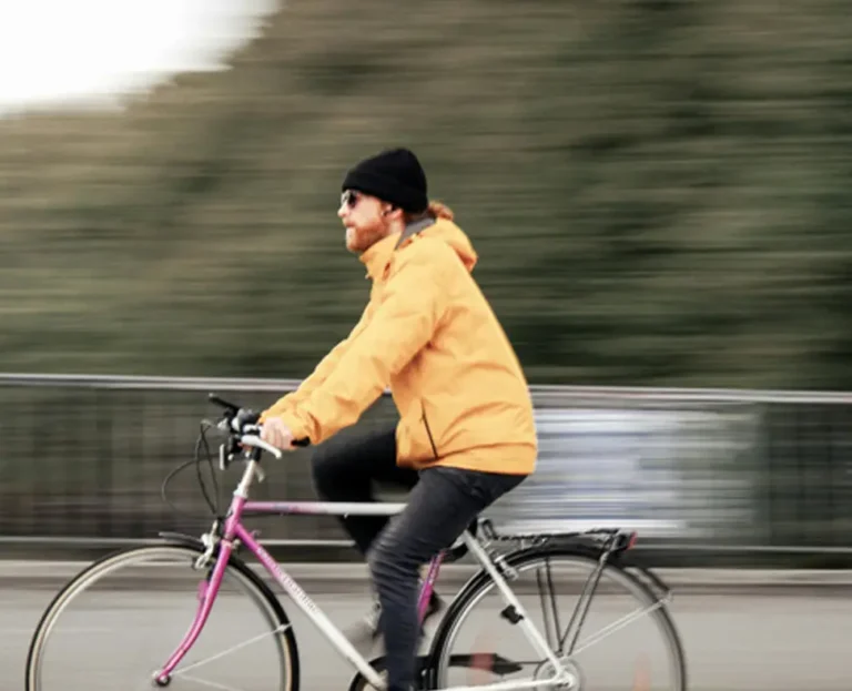 Pessoa andando de bicicleta em uma estrada ou ponte, usando casaco amarelo, calça escura e gorro preto, com fundo desfocado sugerindo movimento.